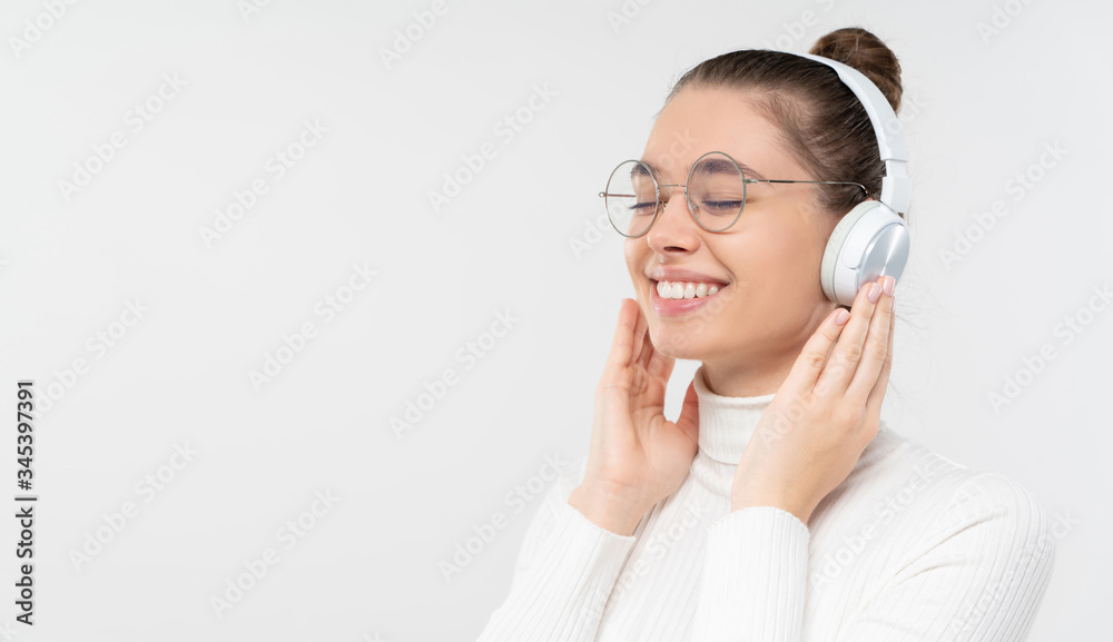 Horizontal banner of young smiling female in glasses, enjoying music through wireless headphones, eyes closed with pleasure, isolated on gray background with copy space Horizontal banner of young smiling female in glasses, enjoying music through wireless headphones, eyes closed with pleasure, isolated on gray background with copy space