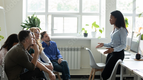 Serious african american female leader, mentor holding briefing for team interns in office, teaching students. Confident diverse businesswoman at company meeting with diverse colleagues.