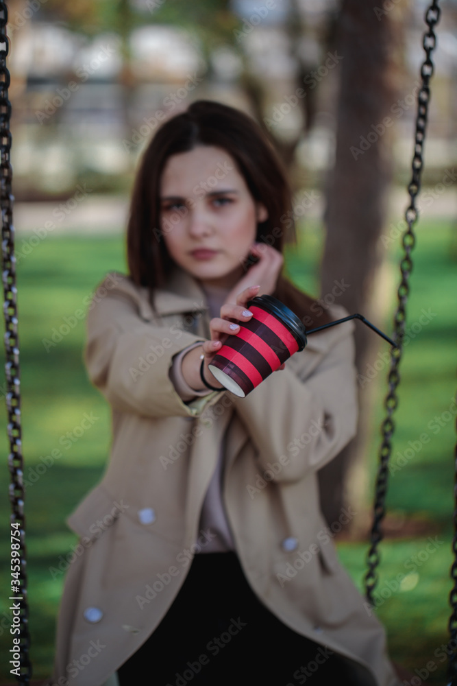 Obraz premium Portrait of a girl with a blurred face, holding a red-black glass with a straw in front of her hands. A disposable glass with a drink in female hands.