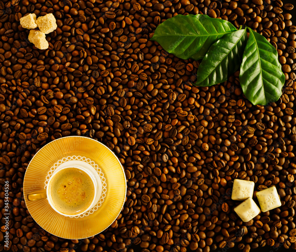 Naklejka premium Roasted coffee beans background white cup on golden saucer with foamy espresso, top view. On beans fresh green coffee leaves, brown sugar cubes, slices of chocolate.