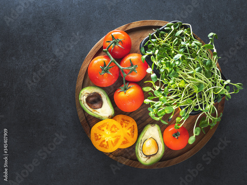 overhead view to red tomatoes with sprouts and avocadon on wooden plate with copy space on dark background