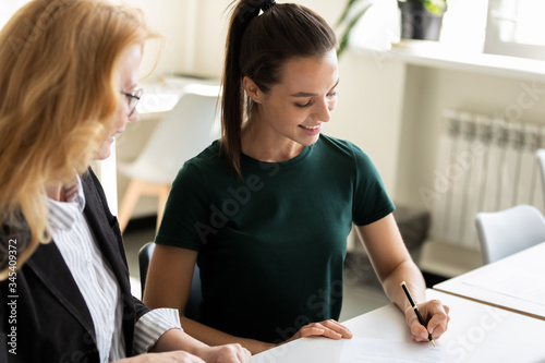 Happy young attractive businesswoman writing signing paper document with mature female partner. Smiling woman puts signature on business document, agreement in office. photo