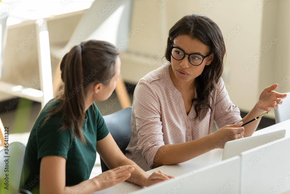 Fototapeta premium Serious young african american businesswoman talking with female leader in boardroom at meeting. Confident diverse mentor with glasses presenting new business concept with woman colleague discuss.