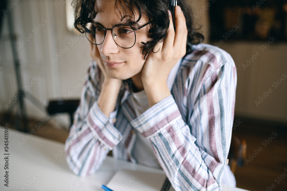 © Anatoliy Karlyuk - Handsome young male in eyewear and casual clothes holding hands on headphones, listening to lecture or podcast online using electronic gadget, learning from home. People, technology and education © Anatoliy Karlyuk - Handsome young male in eyewear and casual clothes holding hands on headphones, listening to lecture or podcast online using electronic gadget, learning from home. People, technology and education