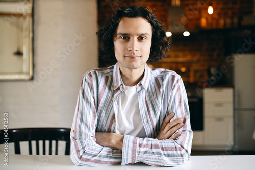 Horizontal image of cool stylish young Caucasian male with black curls relaxing indoors sitting at white desk keeping arms folded, his look and posture expressing confidence, smiling at camera