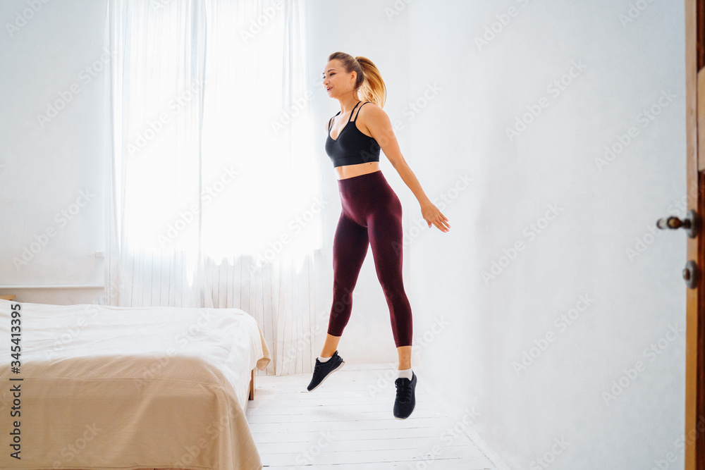 Strong Athletic Fit girl in top and leggings is Energetically Jogging and jumping with smile in Place at Home in Her Spacious and Bright bedroom with Minimalistic white interior. Strong Athletic Fit girl in top and leggings is Energetically Jogging and jumping with smile in Place at Home in Her Spacious and Bright bedroom with Minimalistic white interior.