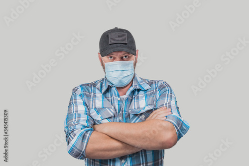 Caucasian man dressed casual with plain blue shirt and black baseball cap wears face medical hat and looks angrily at the camera. Mean looking man with crossed arms and safety mask isolated on gray. 
