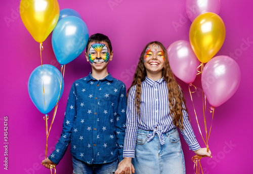 Sister and brother with aquagrim holding colorful inflatable balloons isolated on violet background. Birthday concept