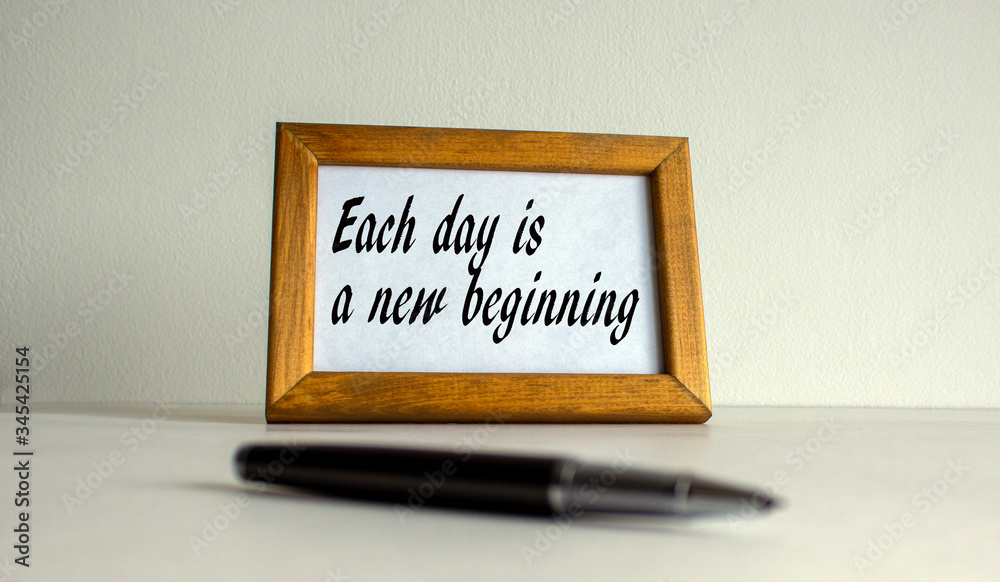 Wooden picture frame with inscription 'each day is a new beginning' on beautiful white fon. Pen on the table. Wooden picture frame with inscription 'each day is a new beginning' on beautiful white fon. Pen on the table.