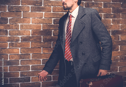 Portrait of fashionable well dressed man with beard posing outdoors looking away, confident and focused mature man in coat standing with suitcase on a brick wall background, elegant fashion model. photo