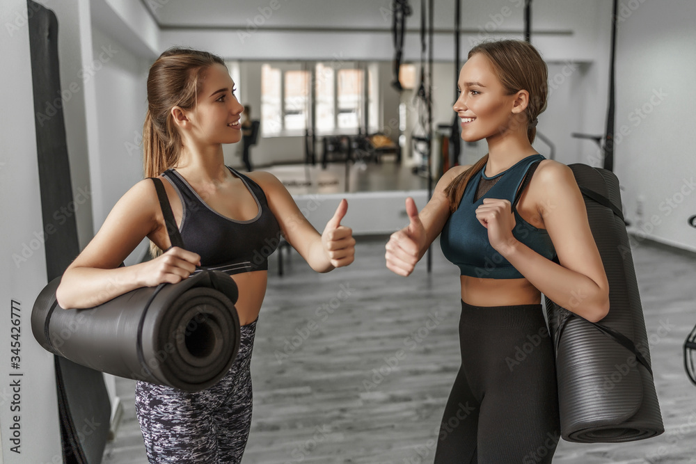 Happy blonde fitness girls, with a yoga mat showing okay signal after team training at modern gym Happy blonde fitness girls, with a yoga mat showing okay signal after team training at modern gym