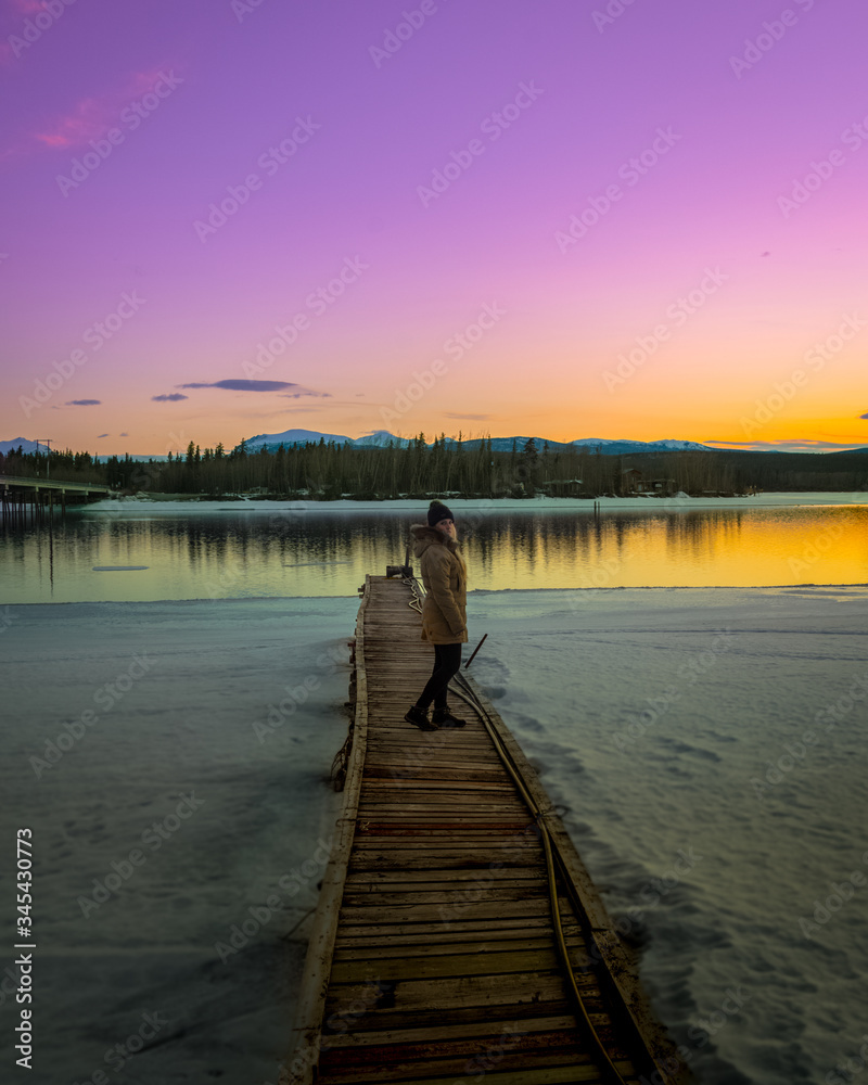 Fototapeta premium Woman standing on a pier, boat launch with snow & purple, orange and pink stunning sunset with mountains and river, lake reflection. 