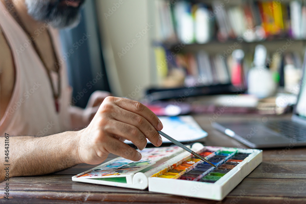 man artist painting with paint brush and watercolor palette on paper in front of laptop, closeup. Selective focus man artist painting with paint brush and watercolor palette on paper in front of laptop, closeup. Selective focus