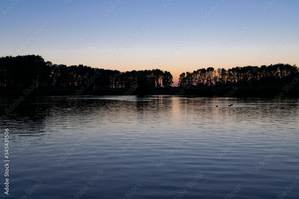 The surface of the water with ripples and reflections of sunset on background of a calm symmetrical landscape in a light misty haze, soft focus.