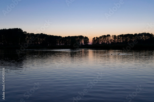 The surface of the water with ripples and reflections of sunset on background of a calm symmetrical landscape in a light misty haze, soft focus.