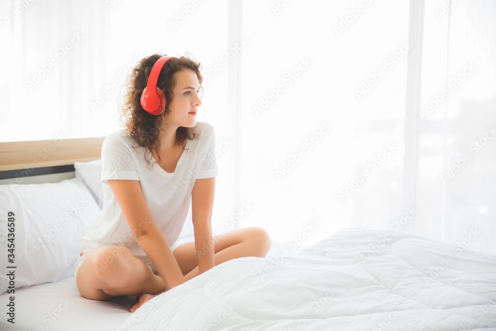 A beautiful woman is wearing a casual white shirt. She is smiling and wearing a red wireless headphones. She is sitting in the back bed and is getting out of bed in the morning. A beautiful woman is wearing a casual white shirt. She is smiling and wearing a red wireless headphones. She is sitting in the back bed and is getting out of bed in the morning.