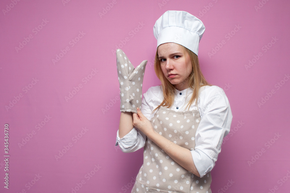 Portrait of a serious cook girl in uniform, apron and gloves for baking on a colored background, beautiful woman housewife in kitchen clothes puts on baking gloves Portrait of a serious cook girl in uniform, apron and gloves for baking on a colored background, beautiful woman housewife in kitchen clothes puts on baking gloves