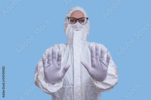 A woman wearing a jumpsuit, goggles, protective mask and white gloves raises her hands in a stop sign.