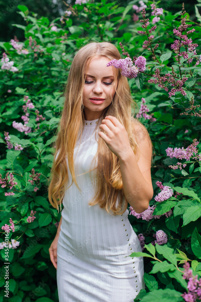 Portrait of a charming blond woman wearing beautiful white dress standing next to lilac bush.