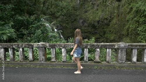 Tracking a young woman walking along the Hana highway, looking out across lush jungle towards the Upper Waikani Falls photo