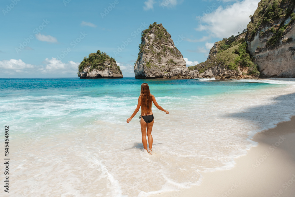 Back view young slim woman weaing black bikini  with long dark hair  on a tropical beach enjoy summer vacation. Vacation in Nusa Penida