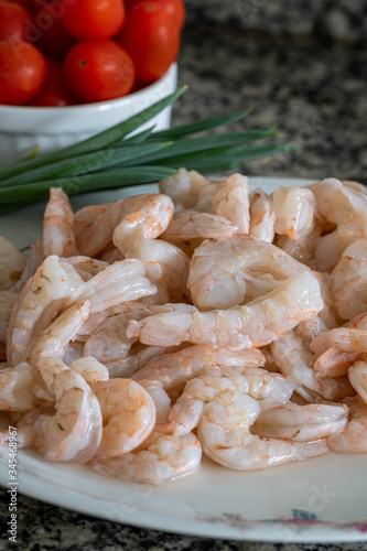 Raw prawns on top of a white plate accompanied by spices such as chives and tomatoes, on top of a gray granite countertop