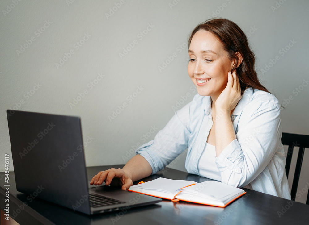 Adult smiling brunette woman doing notes in daily planner with opened laptop at the table Adult smiling brunette woman doing notes in daily planner with opened laptop at the table