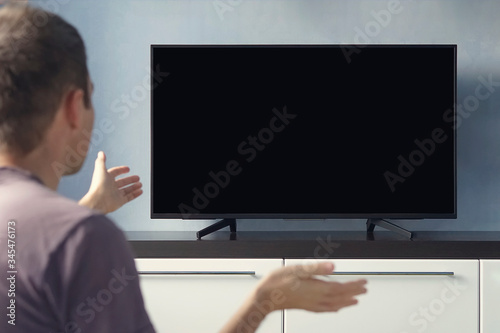 A young guy emotionally watching TV and waving his hands in discontent. Blank screen for design. Frustrated Man Sitting On Sofa In Front Of Television With No Signal. photo