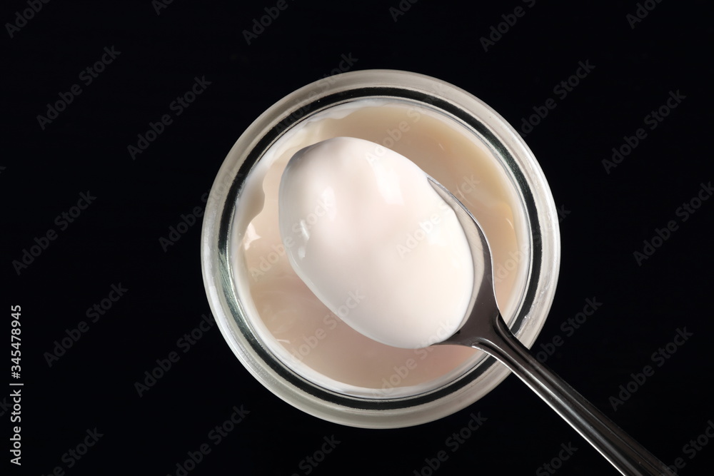 Spoon with yogurt on open small glass jar at black background. D Spoon with yogurt on open small glass jar at black background. D