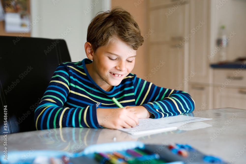 Hard-working happy school kid boy making homework during quarantine time from corona pandemic disease. Healthy child writing with pen, staying at home. Homeschooling concept Hard-working happy school kid boy making homework during quarantine time from corona pandemic disease. Healthy child writing with pen, staying at home. Homeschooling concept