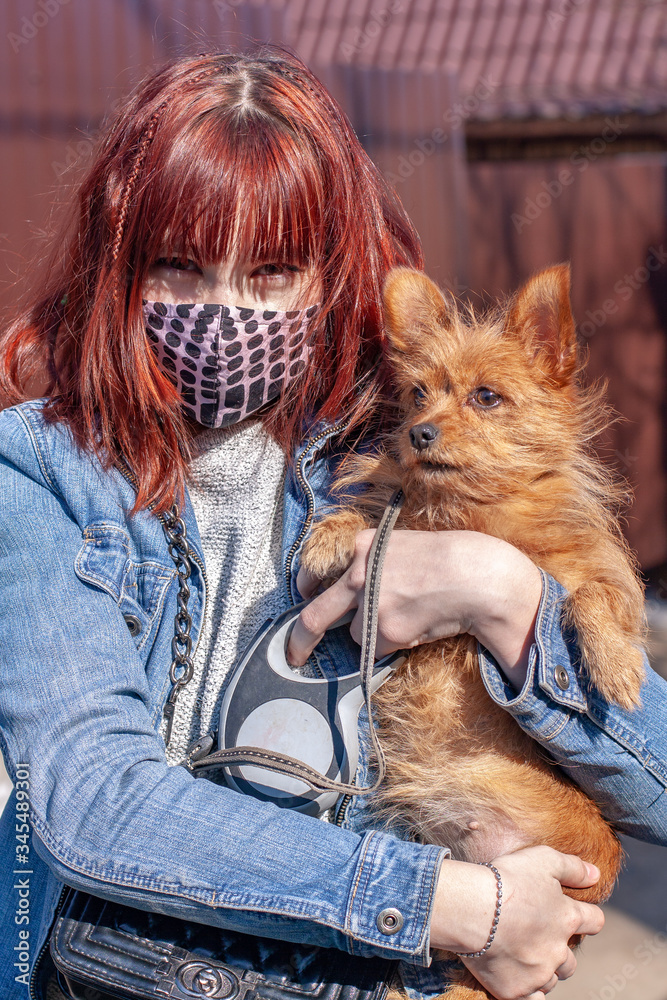 The red-haired girl in a homemade protective mask holds a funny little dog of the Yorkshire Terrier on the street. Dog walking during coronavirus covid-19 and quarantine. Brown dog hair. Vertical. The red-haired girl in a homemade protective mask holds a funny little dog of the Yorkshire Terrier on the street. Dog walking during coronavirus covid-19 and quarantine. Brown dog hair. Vertical.