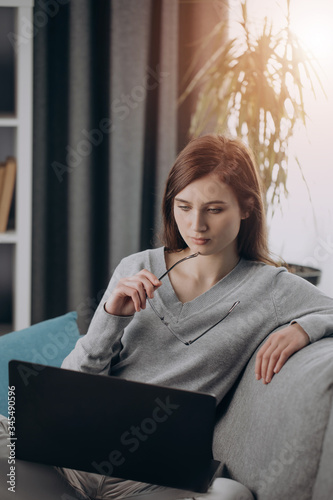 Thoughtful woman with dark hair sitting on cozy sofa and looking at computer screen. Young girl in grey sweater and white trousers working at home on laptop