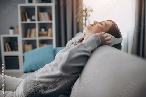 Side view of beautiful lady enjoying leisure time at home with favorite music. Relaxed woman in grey couch and wireless headphones sitting on couch.