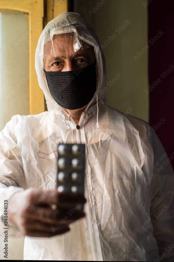 Senior man with corona preventive mask and PPE suit showing medicine strip in front of a window. Covid, Lock down and Home isolation