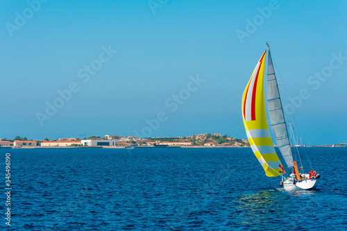 A view of a colorful lonely sailboat as it sails tilted by the wind in the Mediterranean sea with the coast in the background on a sunny day with the blue sky on summer, in Sardinia Italy 