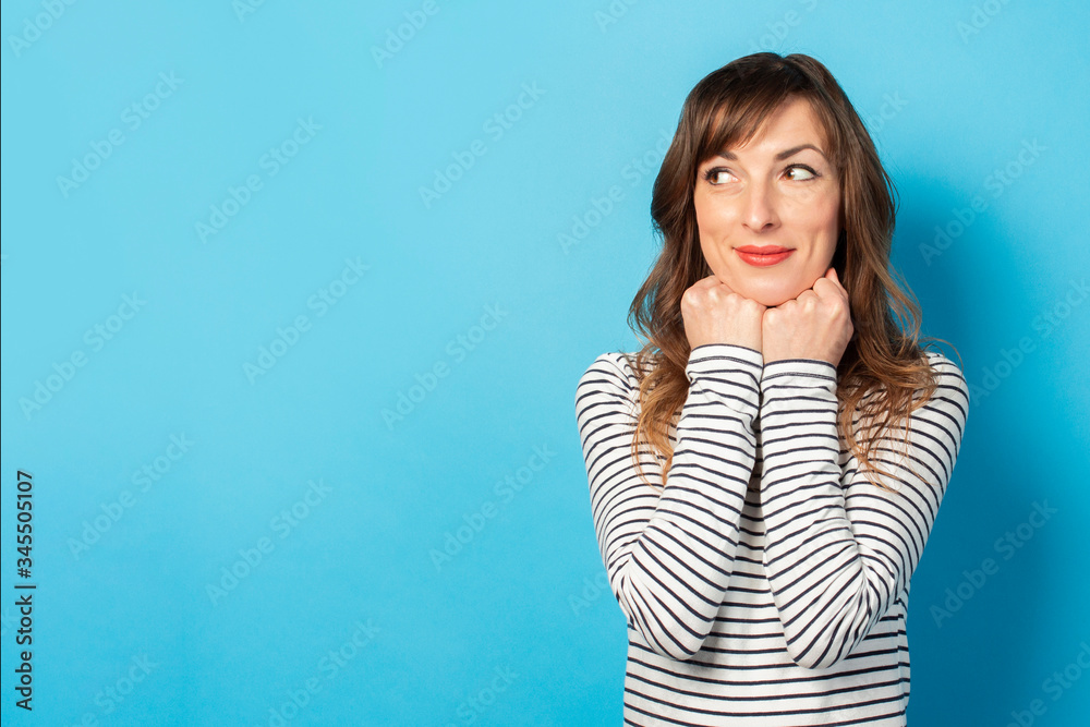 Portrait of a young friendly woman with a smile in a casual t-shirt holds her hands under her chin and looks away on an isolated blue background. Emotional face. Gesture rejoices in something Portrait of a young friendly woman with a smile in a casual t-shirt holds her hands under her chin and looks away on an isolated blue background. Emotional face. Gesture rejoices in something