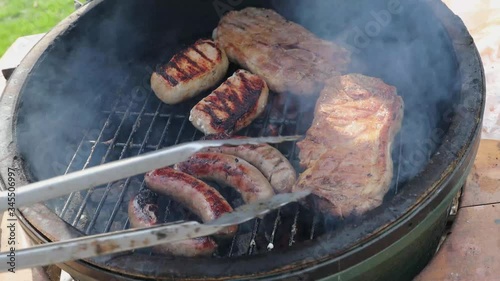 A kamado style grill full of steaks and brats with tongs moving the meat around photo