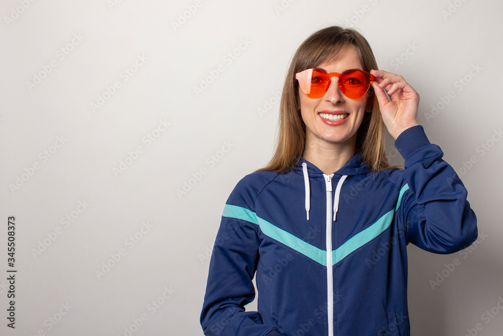 smiling young woman in a 90s sports jacket and in red glasses on a light background. Model holds the edge of the glasses with his hand. Concept of style, fashion, sport smiling young woman in a 90s sports jacket and in red glasses on a light background. Model holds the edge of the glasses with his hand. Concept of style, fashion, sport