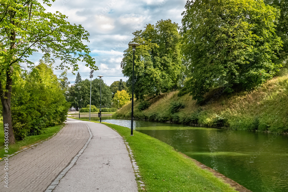 A pond in Snelli park, Tallinn, Estonia. Green trees and sidewalk on summer day with clouds.