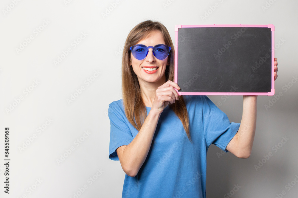 Portrait of a young friendly woman with a smile in a casual blue t-shirt, blue glasses, holding a black chalkboard with blank space for text on an isolated light background. Emotional face