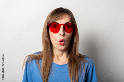 Portrait of a young friendly woman with a surprised face in a casual blue t-shirt and red glasses on an isolated light background. Emotional face