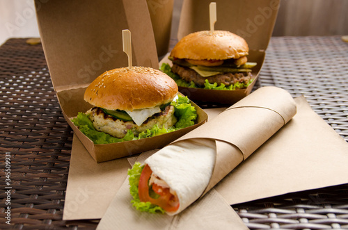 two different burgers in boxes and chicken roll with vegetables on a paper sheet