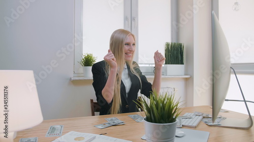Elegant blond female sitting at desk at workplace with computer holding in hands bundle of cash looking astonished at monitor, and throwing money in air and happily making yes gesture.