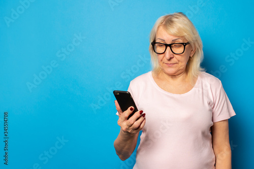 Portrait of an old friendly woman in glasses and a casual t-shirt holding a phone in her hands and looking at the screen on an isolated blue background. Emotional face