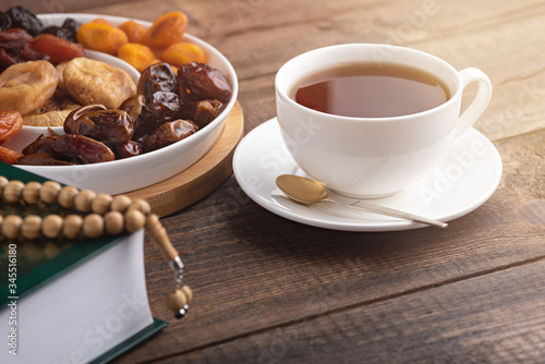 iftar concept, white Cup of tea, plate of dried fruit, Koran with prayer beads on brown wooden table in sun