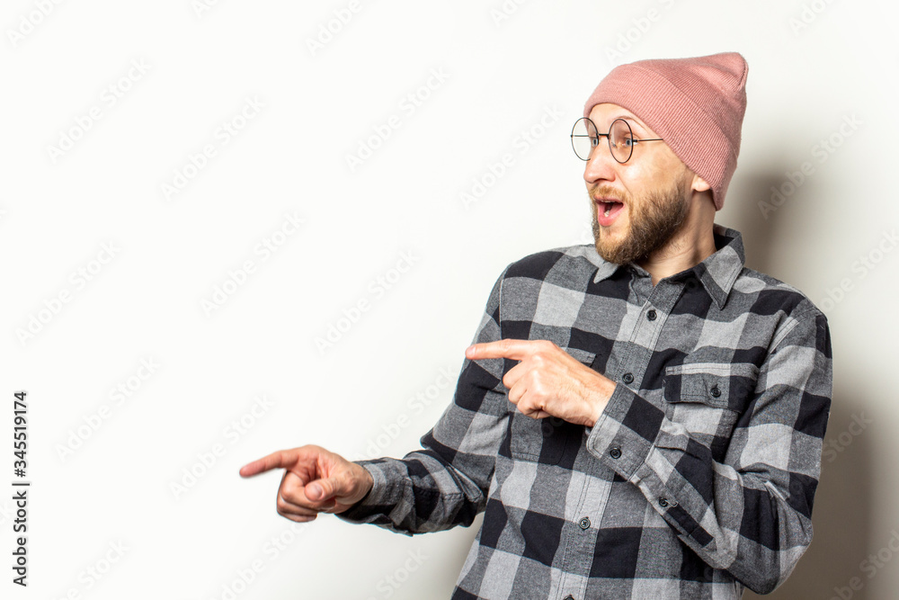Portrait of a young man with a beard in a hat, a plaid shirt and glasses points his fingers to the side with a surprised face on an isolated light background. Emotional face. Gesture look at it Portrait of a young man with a beard in a hat, a plaid shirt and glasses points his fingers to the side with a surprised face on an isolated light background. Emotional face. Gesture look at it