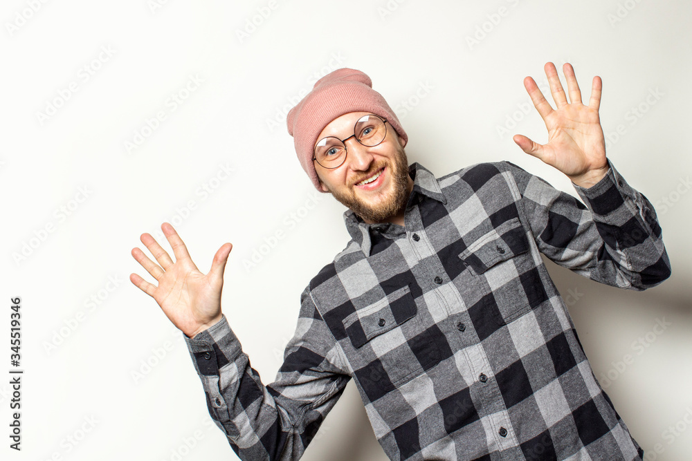 Naklejka premium Portrait of a young man with a beard in a hat, a plaid shirt peeps from behind the edge of the picture on an isolated light background. Emotional face. Gesture surprise shock