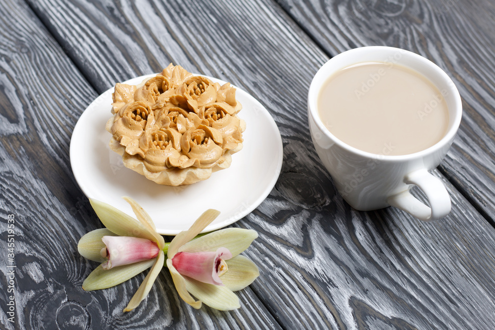 Cupcake and a cup of coffee with milk. Nearby are orchid flowers. Cake decorated with flowers made of butter cream. The cream has a caramel color. On brushed pine boards painted black.