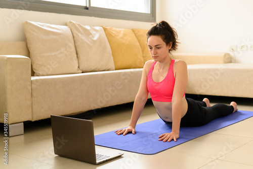 Young athletic girl does yoga on a sports blue rug in sportswear. A laptop with online lessons, she looks at the screen and repeats the exercises. Online life concept. Healthy lifestyle. Copy space.
