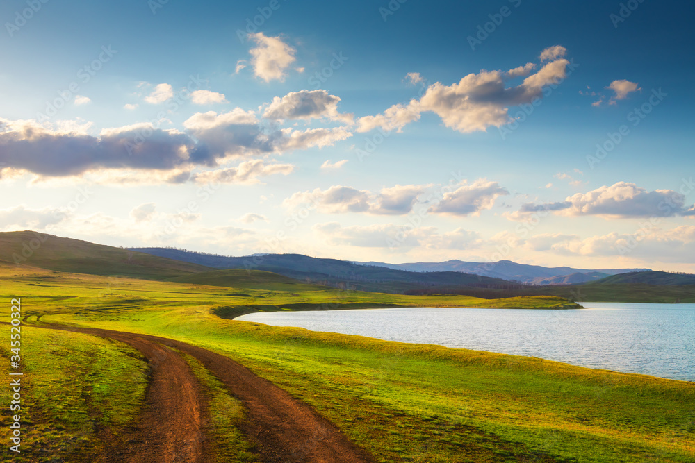Road near the lake in the mountains. Spring nature landscape at sunset. Fresh green grass on the hills. South Ural, Bashkortostan Republic, Russia.